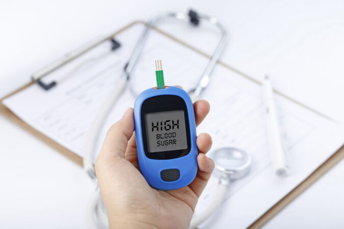 Hand holding a blood glucose meter measuring blood sugar, the background is a stethoscope and chart file