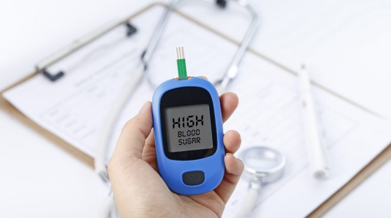 Hand holding a blood glucose meter measuring blood sugar, the background is a stethoscope and chart file