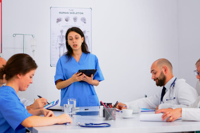 Medical people meeting and planning with shareholders in hospital office sitting at desk. Doctors and nurses brainstorm ideas together, physicians diagnosis and presentation data using tablet