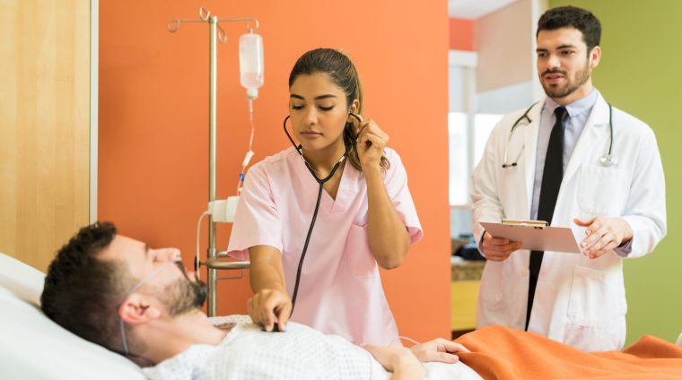 Male And Female Doctor Treating Patient At Hospital Female doctor examining sick patient with stethoscope while male doctor analyzing reports at hospital