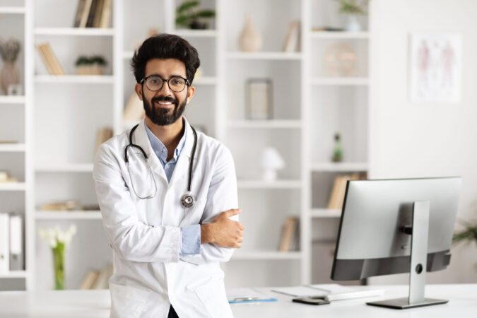 Happy handsome young Indian man doctor, standing confidently next to a table with computer, poses in a well-equipped clinic, symbolizing modern healthcare professionalism, copy space