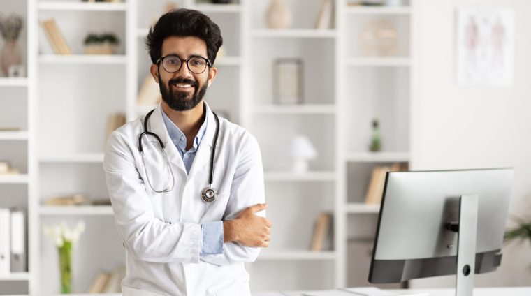 Indian Doctor Poses Beside Computer in Clinic Setting Happy handsome young Indian man doctor, standing confidently next to a table with computer, poses in a well-equipped clinic, symbolizing modern healthcare professionalism, copy space