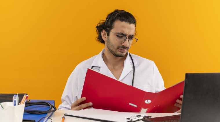 thinking young male doctor with medical glasses wearing medical robe with stethoscope sitting at desk work on laptop with medical tools holding and looking folder on isolated yellow background with copy space online fellowship courses after mbbs, course for doctors, medical cert online,
